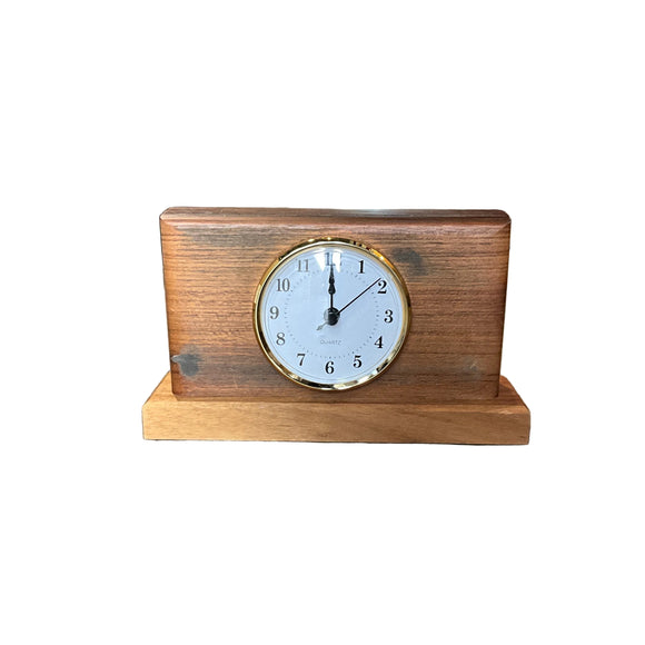 A small wooden desk clock with a rectangular wood body and a round white clock face, photographed on a concrete floor in a cluttered indoor shop or warehouse setting.