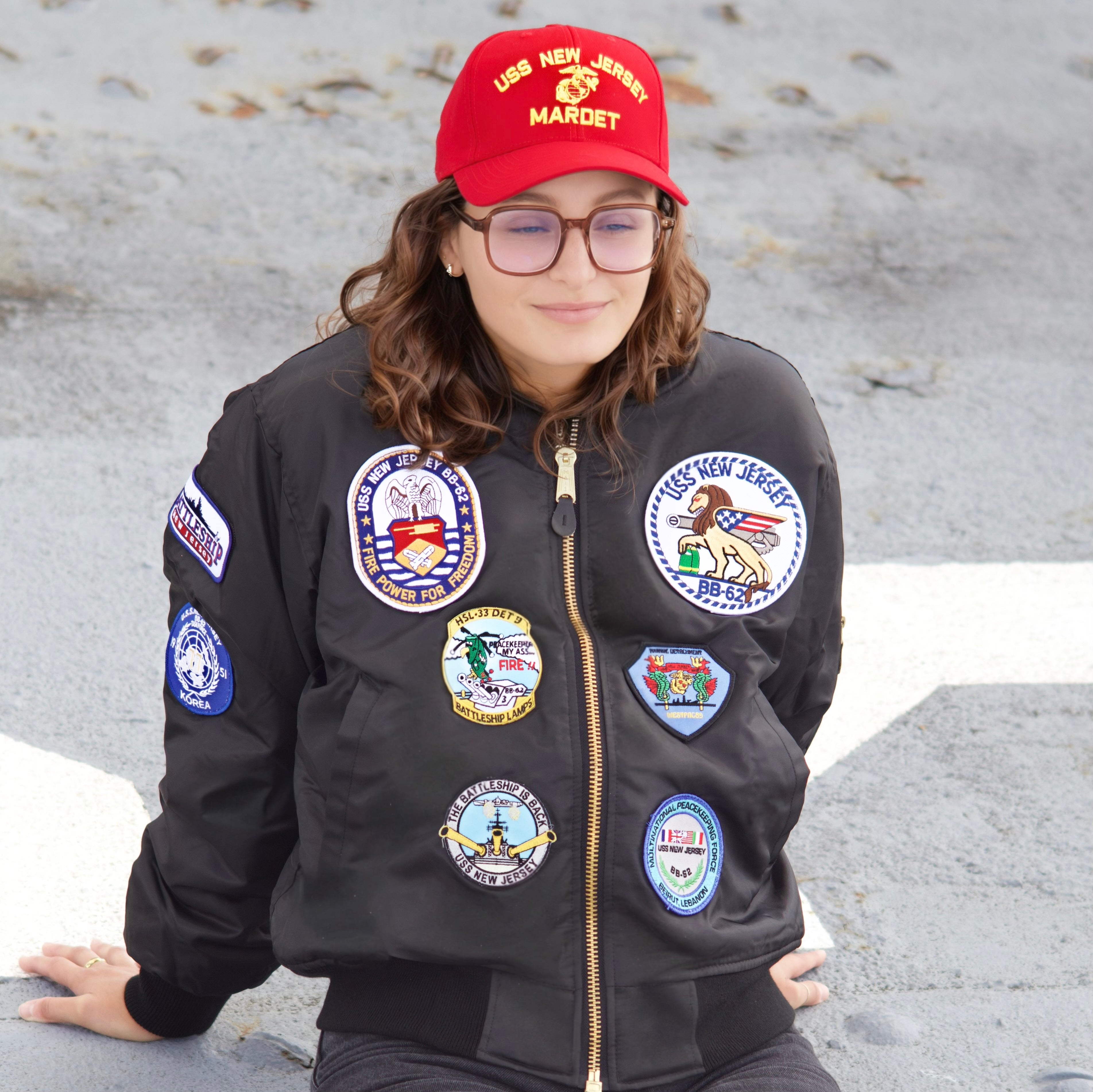 Emma Hermo wearing a battleship New Jersey legacy jacket with patches and a red cap on a stone surface. Commercial Photo Shoot. 